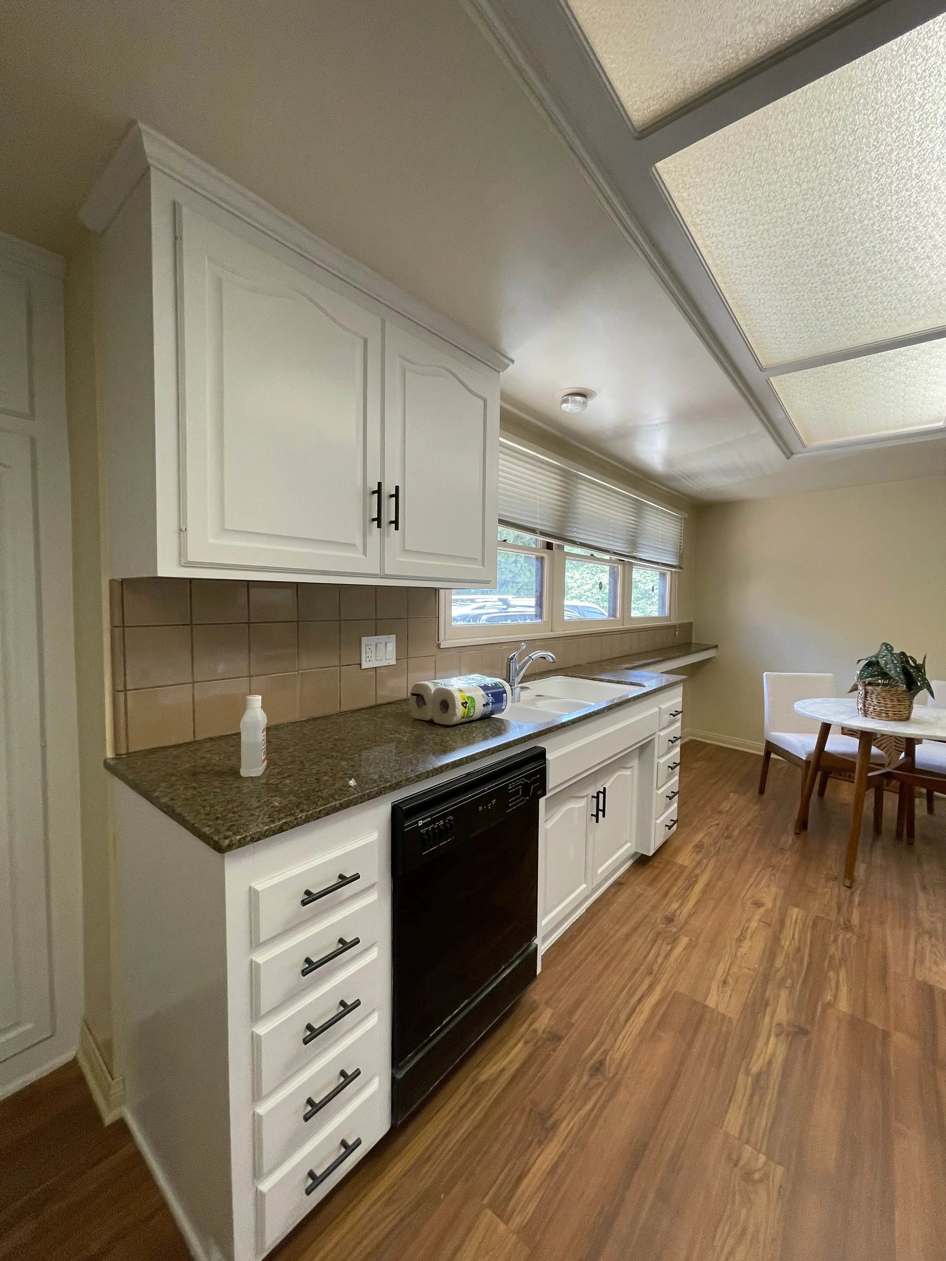 White painted kitchen cabinets with black hardware, granite countertops, brown tile backsplash, and freshly painted ceiling with skylights