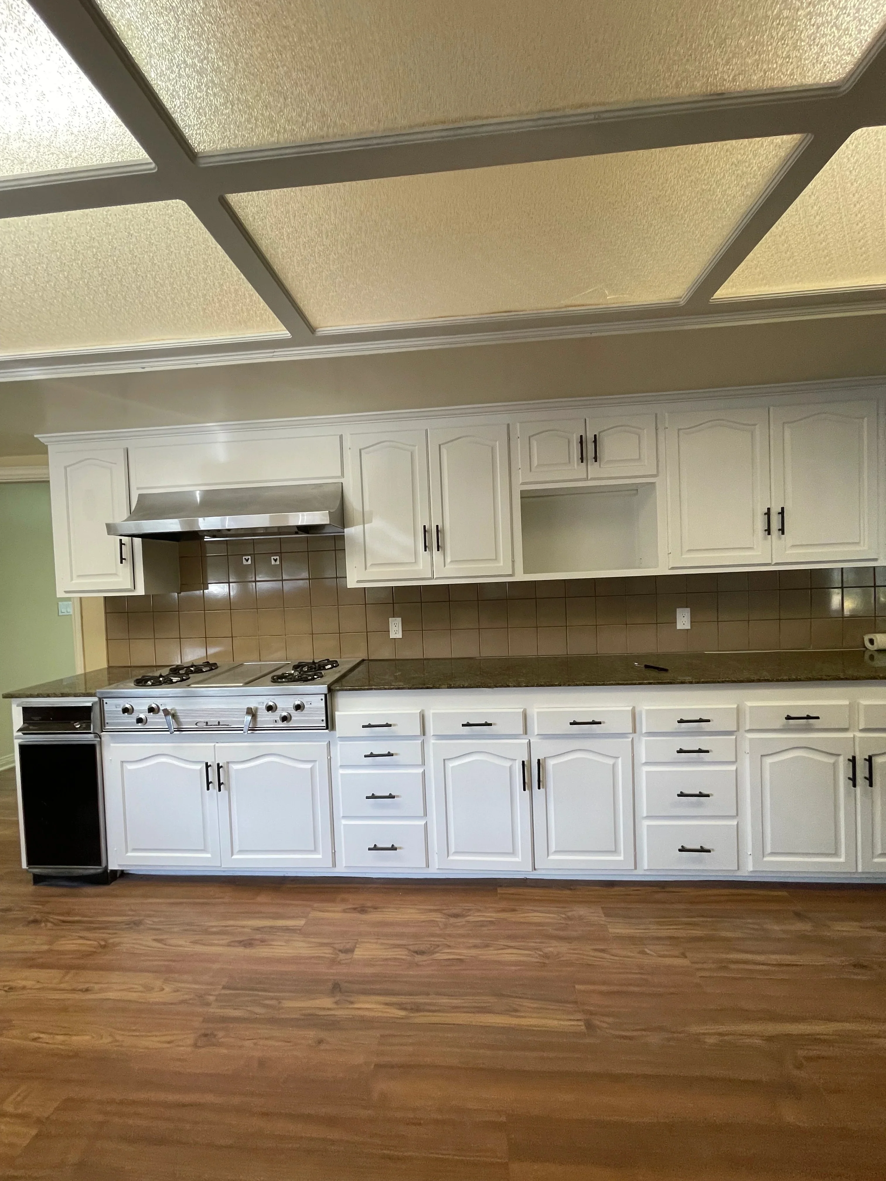 White painted kitchen cabinets with black handles, brown tile backsplash, stainless steel range hood, and coffered ceiling